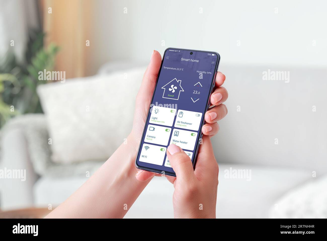 Die Hand einer Frau hält ein Mobiltelefon und zeigt eine Smart-Home-App. Smart Living-Konzept. Dekoriertes Wohnzimmer im Hintergrund Stockfoto