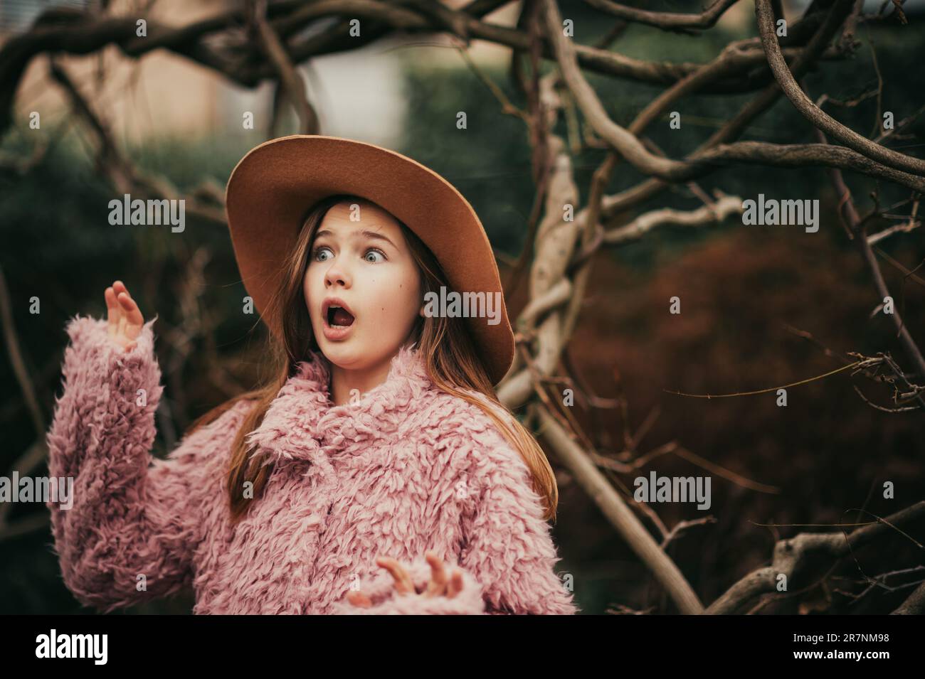 Komisches Mädchen, das vorgibt, Angst zu haben, posiert im dunklen Wald, trägt braunen Hut und rosa Mantel Stockfoto