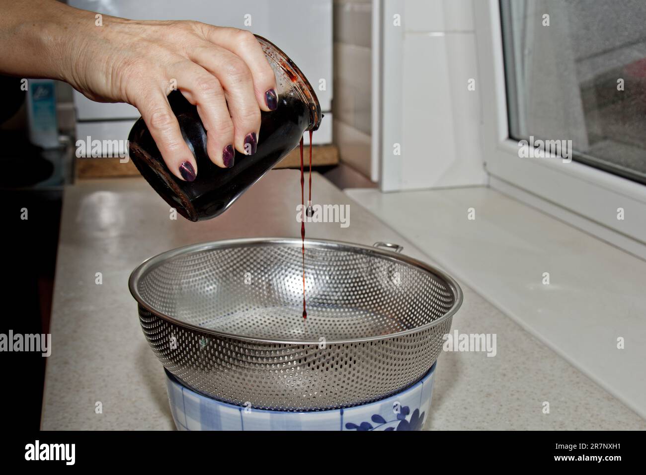 Weibliche Hände gossen Marmelade aus einem Glas durch ein Sieb vor dem Hintergrund des Kücheninnenraums in eine Schüssel. Backen, Kochen Stockfoto