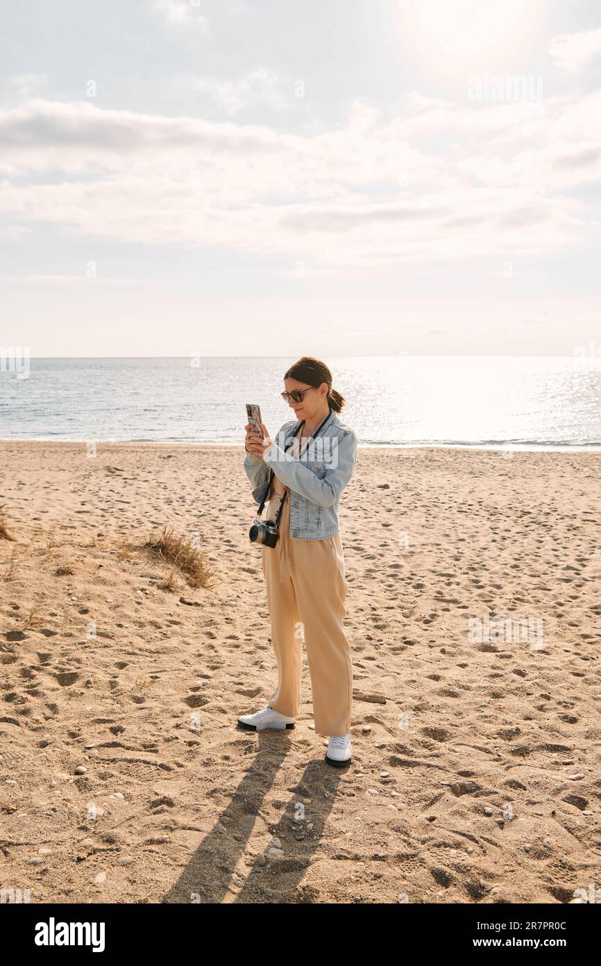 Eine junge, dunkle, reife Frau trägt am Strand bequeme Kleidung und eine Jeansjacke. Sie hat eine alte Kamera um den Hals, während sie sich umhängt Stockfoto