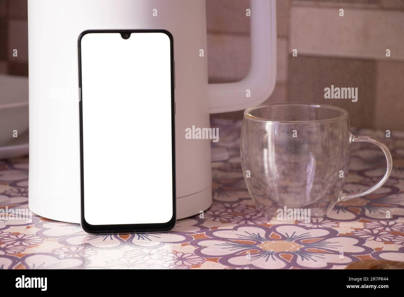 Ein Telefon mit einem leeren weißen Bildschirm steht neben einer weißen Teekanne und einer Tasse auf dem Küchentisch Stockfoto