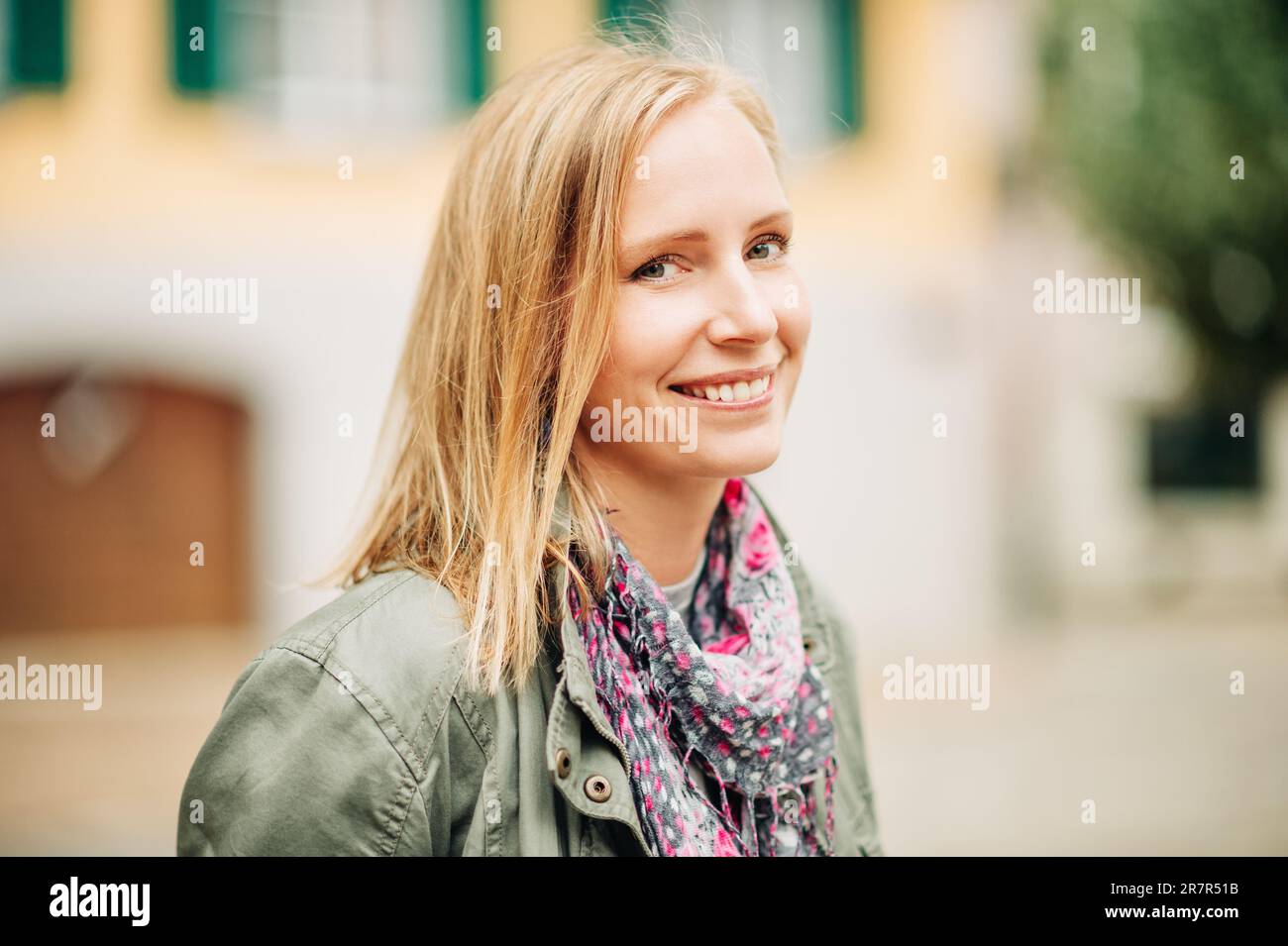 Außenporträt einer wunderschönen blonden Frau, die draußen posiert, einen khaki-Parka und einen rosa Schal trägt Stockfoto