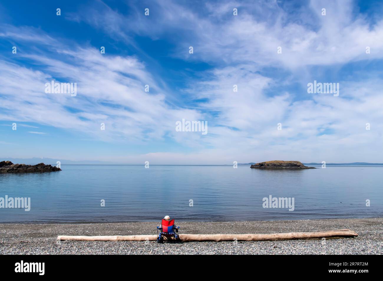 Blick vom Rosario Beach im Deception Pass State Park, WA, USA auf das Wasser der Rosaria Strait mit einem einsamen Mann im Stuhl, der dem Wasser gegen ein Stück Wasser gegenübersteht Stockfoto