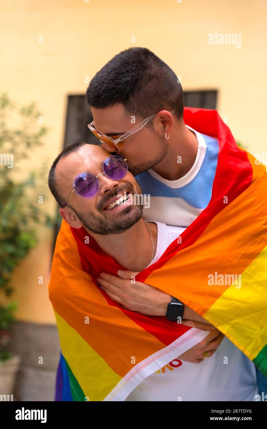 Porträt von zwei homosexuellen Männern, die auf der Spitze sitzen und sich auf der Pride-Party einen kleinen Kuss geben, mit Regenbogenflagge, lgbt-Konzept Stockfoto