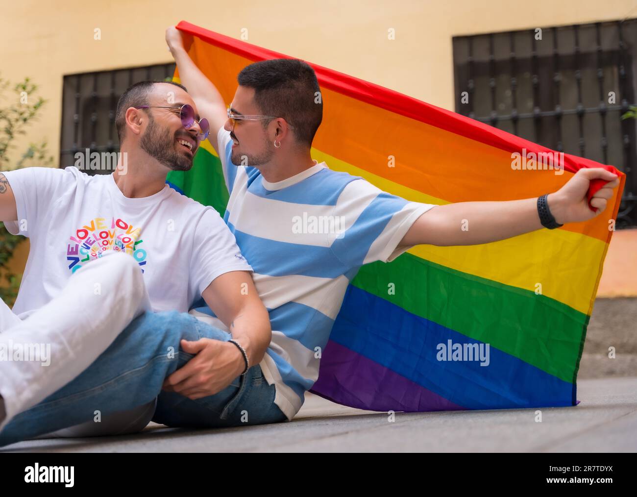 Porträt eines schwulen männlichen Paares, das auf dem Boden sitzt, mit Regenbogenflagge auf der Pride Party, lgbt Concept Stockfoto