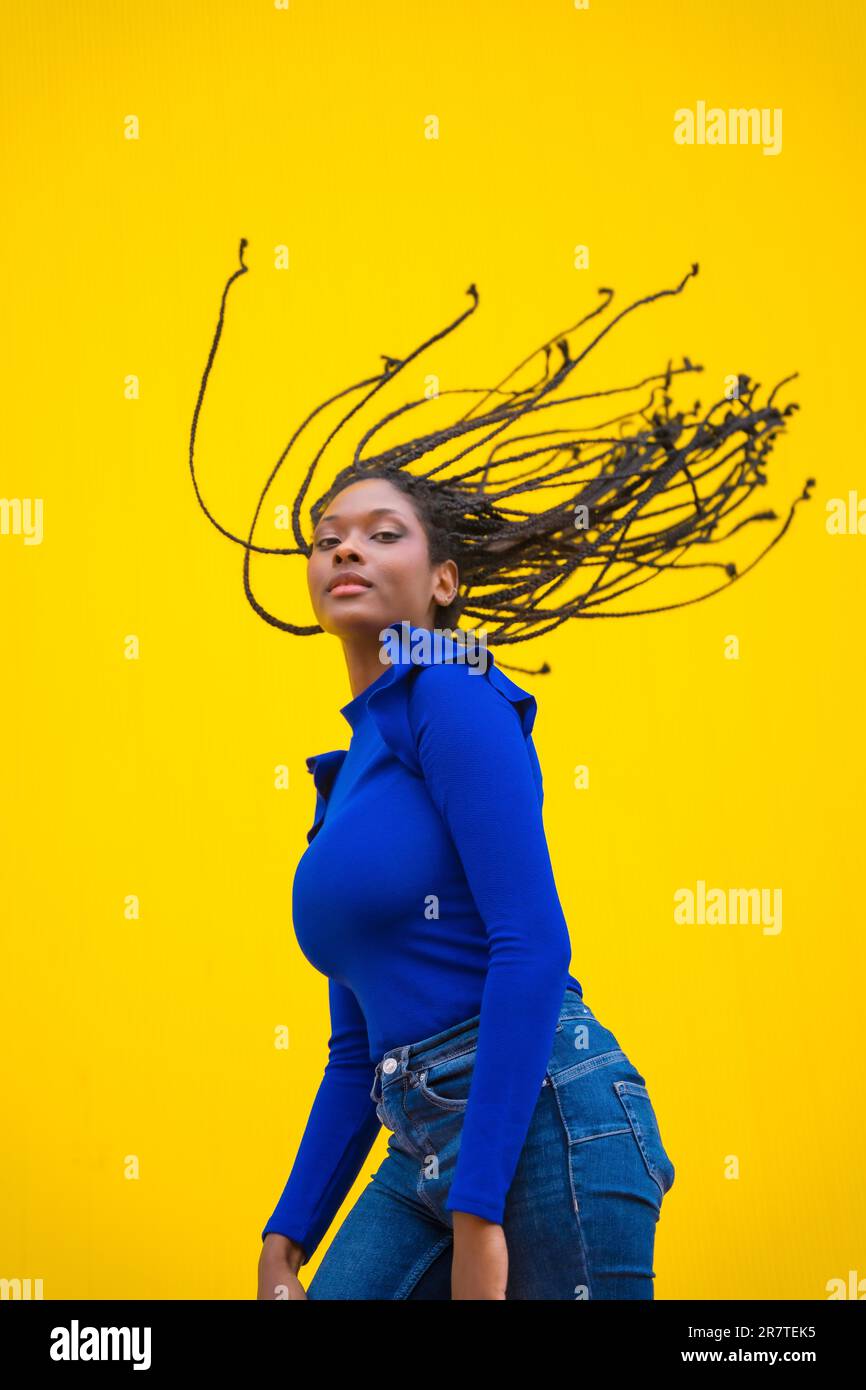 Urbane Sitzung. Junge, dunkelhäutige Frau mit langen Zöpfen in einem blauen Kleid auf gelbem Hintergrund, mit einem ernsten Blick und ihren Zöpfen Stockfoto