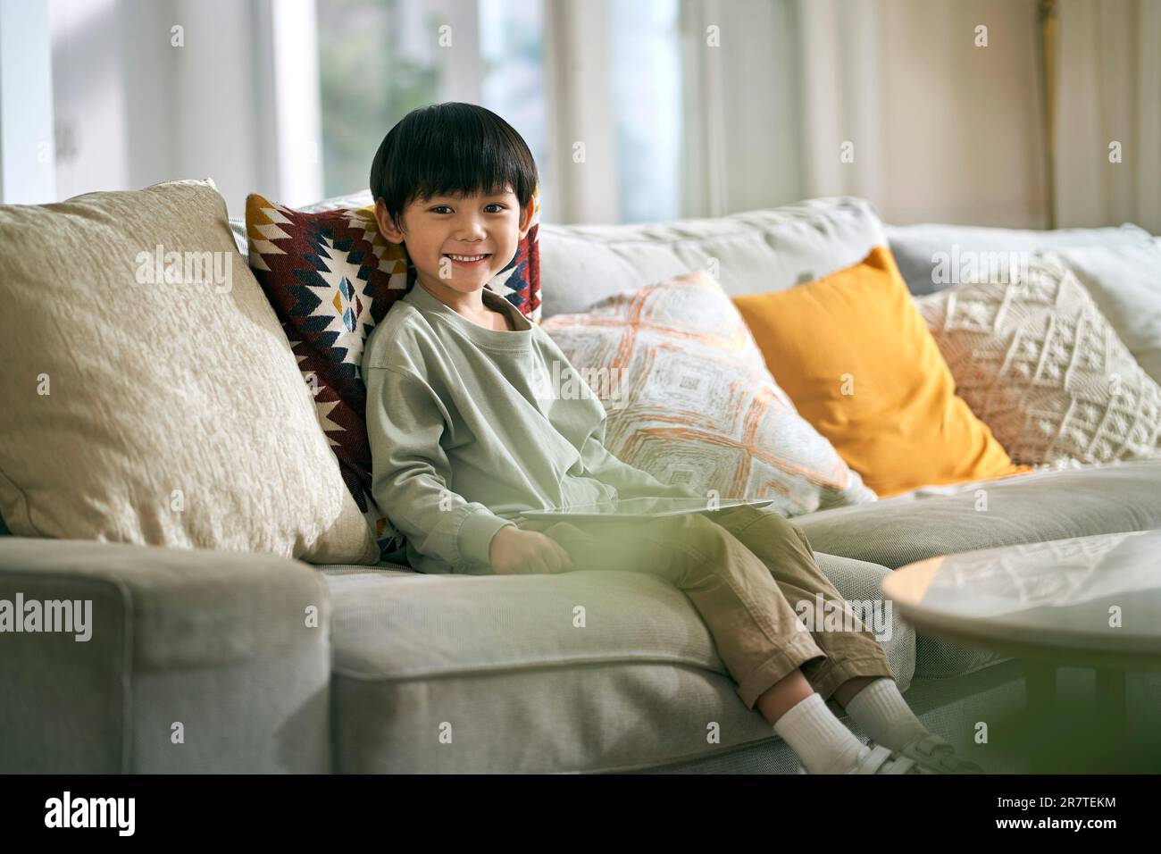 Kleiner asiatischer Junge, der auf der Familientischcouch im Wohnzimmer sitzt und die Kamera ansieht und lächelt Stockfoto