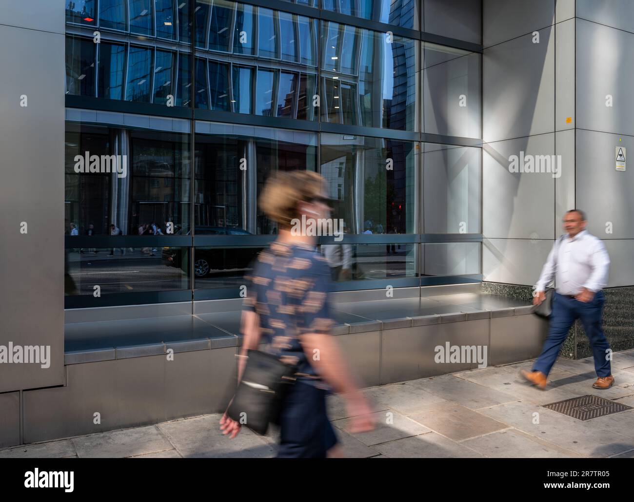 London, Vereinigtes Königreich: Zwei Personen, die mit Bewegungsunschärfe entlang Bishopsgate in der Londoner City spazieren. Die Straßenszene spiegelt sich im Fenster des Gebäudes wider. Stockfoto