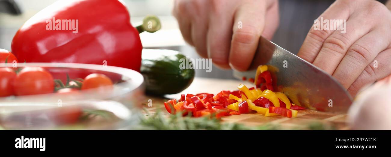 Die Hände des Kochs schneiden gelbe und rote Paprika auf dem Schneidebrett zusammen mit anderem Gemüse auf dem Tisch in der Küche Stockfoto