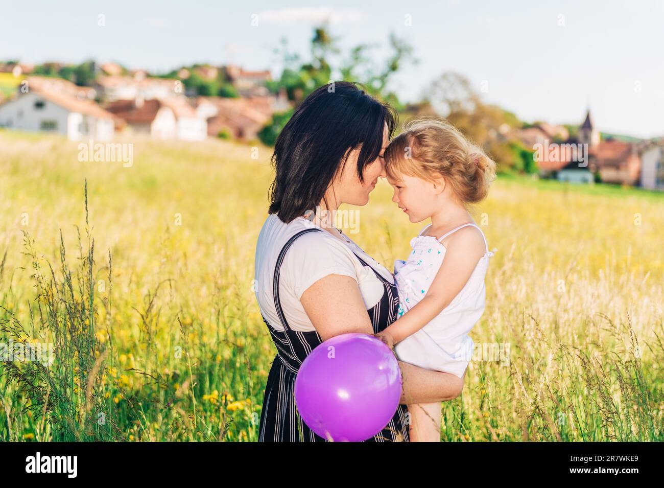 Glückliche junge Mutter, die eine kleine Tochter in den Armen hält, mitten auf einem wunderschönen Sommerfeld, Kind, das mit einem lila Ballon spielt Stockfoto