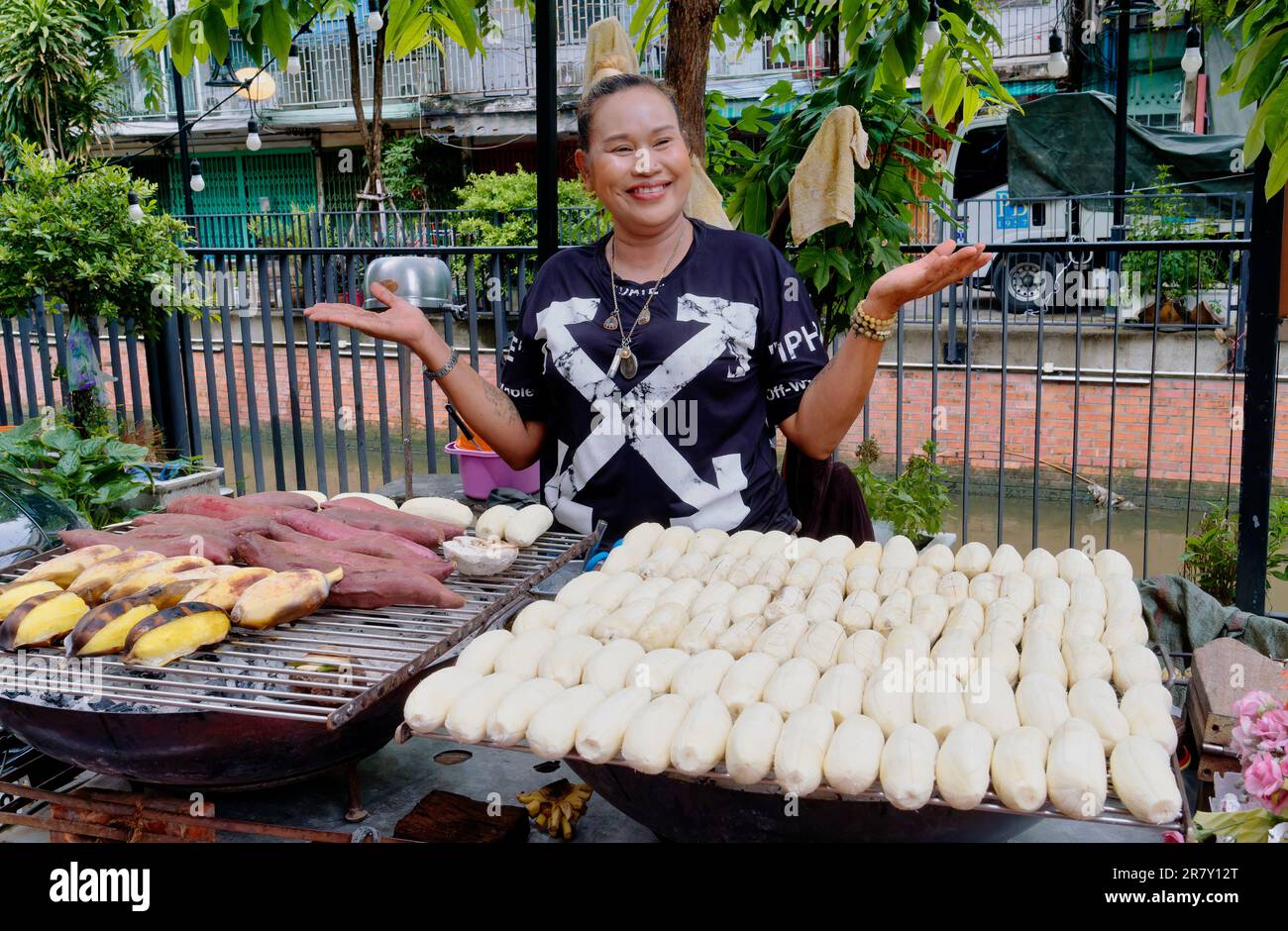 Eine fröhliche thailändische Frau, die einladend an ihrem Straßenstand gestikuliert hat und verschiedene Arten gerösteter Bananen und Manyok oder Kassava verkauft Stockfoto