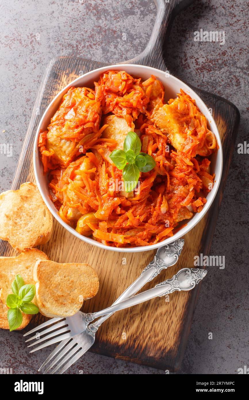 Geschmorter Seehecht mit Tomaten-, Karotten- und Zwiebelnaht auf dem Teller auf dem Holzbrett. Vertikale Draufsicht von oben Stockfoto