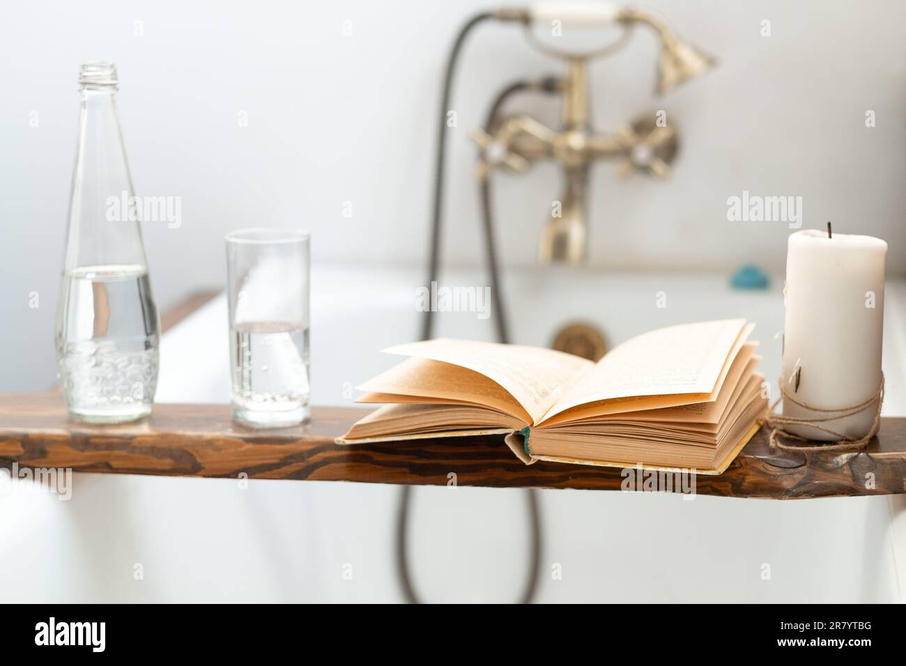 Ein Buch, eine Flasche Wasser, ein Glas und eine Kerze auf einem Holztablett über dem Badezimmer vor dem Hintergrund eines antiken Wasserhahns Stockfoto