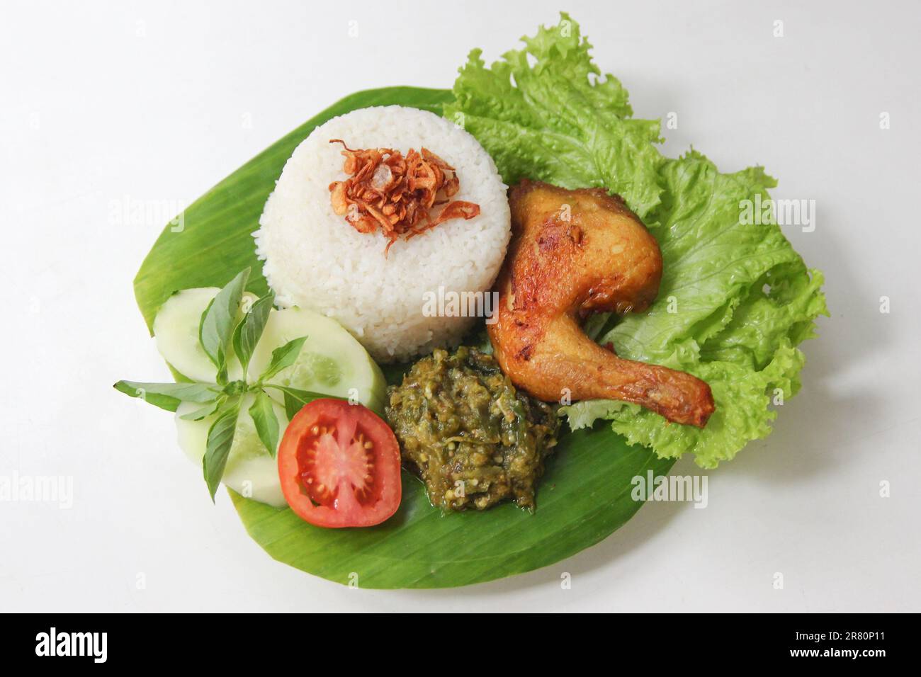 Galangal gebratenes Hähnchen, Reis, Sambal, Tomaten, Gurke, Basilikum, Salat. Indonesische Küche auf Bananenblatt-Teller. Knusprig gebratenes Hähnchen und grüner Sambal Stockfoto