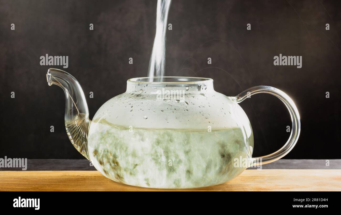 Eine wunderschöne Teekanne aus Glas zum Brühen von Tee steht auf einem Holzbrett auf dunklem Hintergrund, die Teekanne ist transparent. Heißes Wasser wird in den Wasserkocher gegossen Stockfoto
