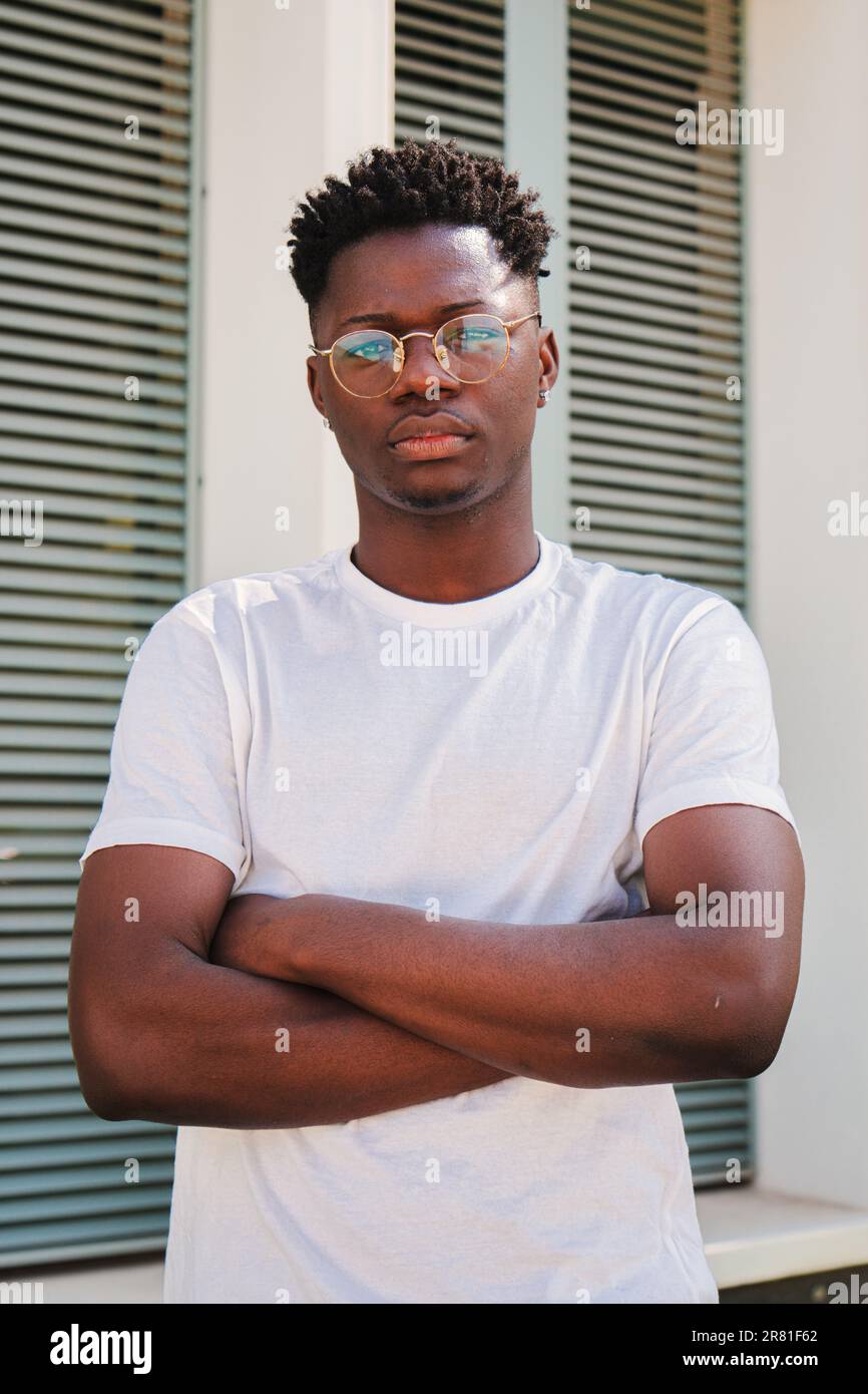 Vertikales Porträt eines jungen afroamerikanischen, selbstbewussten Mannes, der auf die Kamera schaut, mit gekreuzten Armen, die draußen stehen. Ein hübscher Student, der mit einer ruhigen, nachdenklichen Einstellung an der Universität posiert. Hochwertiges Foto Stockfoto