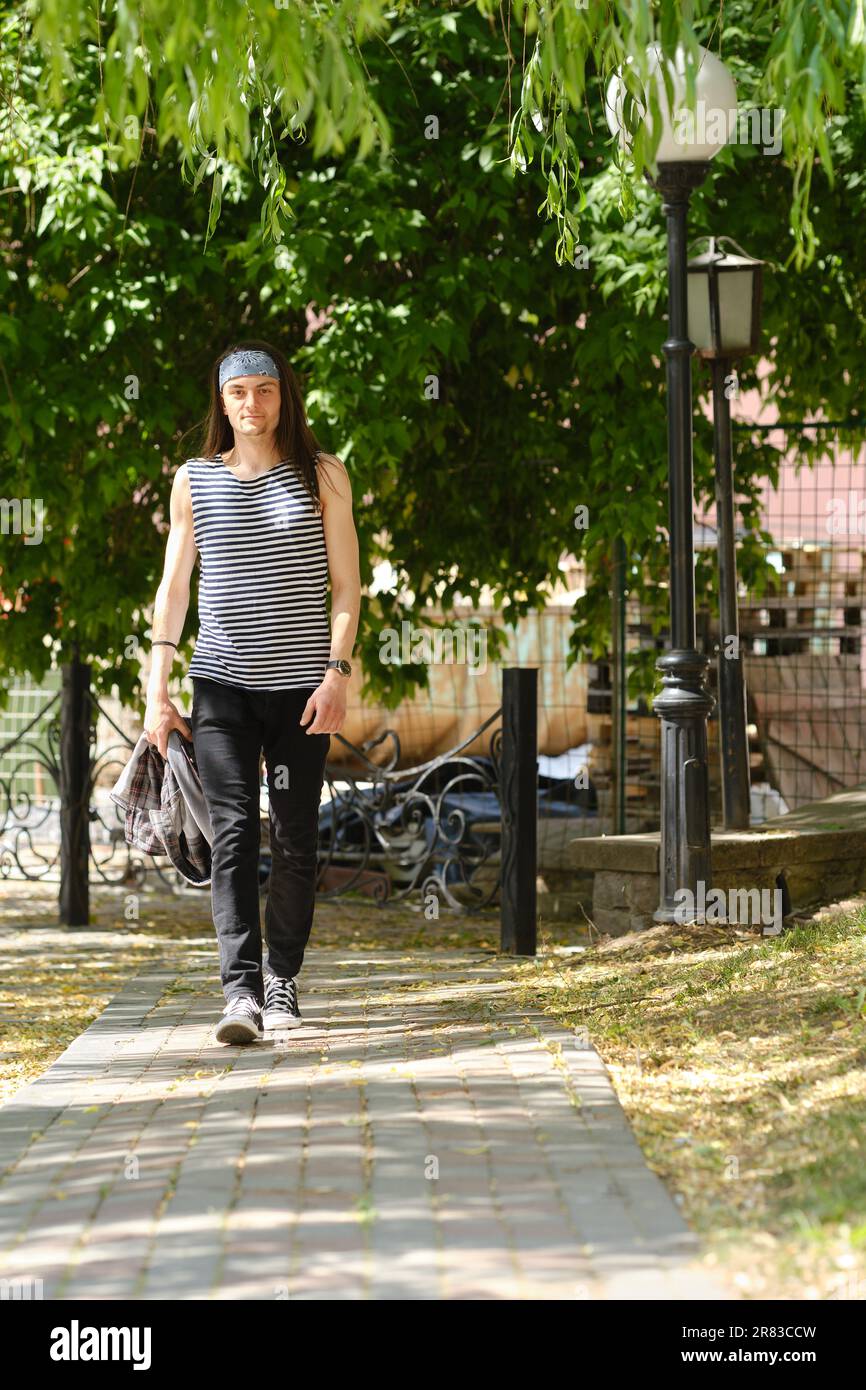 Ein junger Mann mit langen Haaren im Tanktop und Jeans geht auf dem Bürgersteig Stockfoto