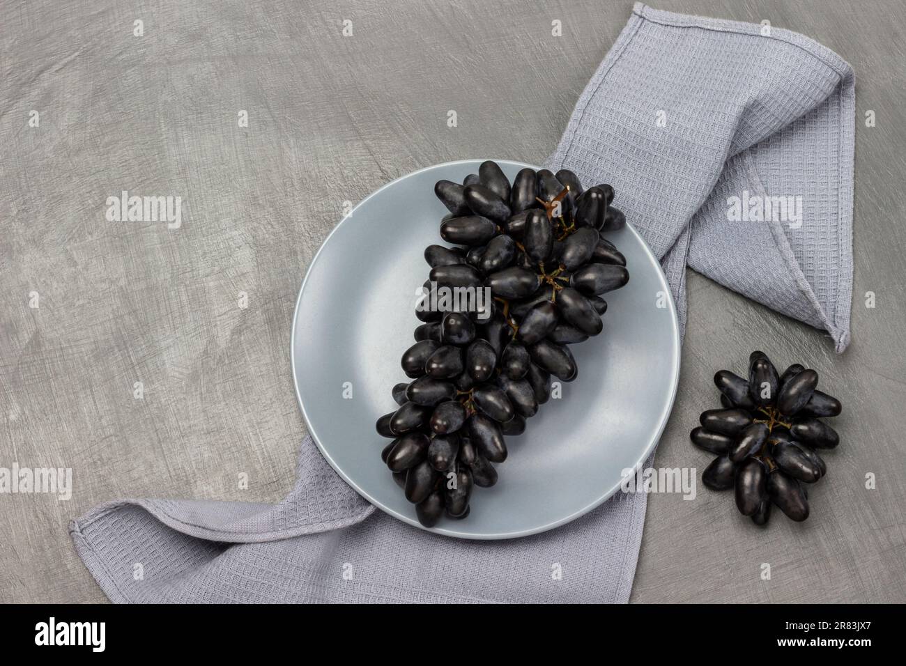 Ein Haufen schwarze Trauben auf einem grauen Teller. Serviette auf dem Tisch. Grauer Hintergrund. Stockfoto