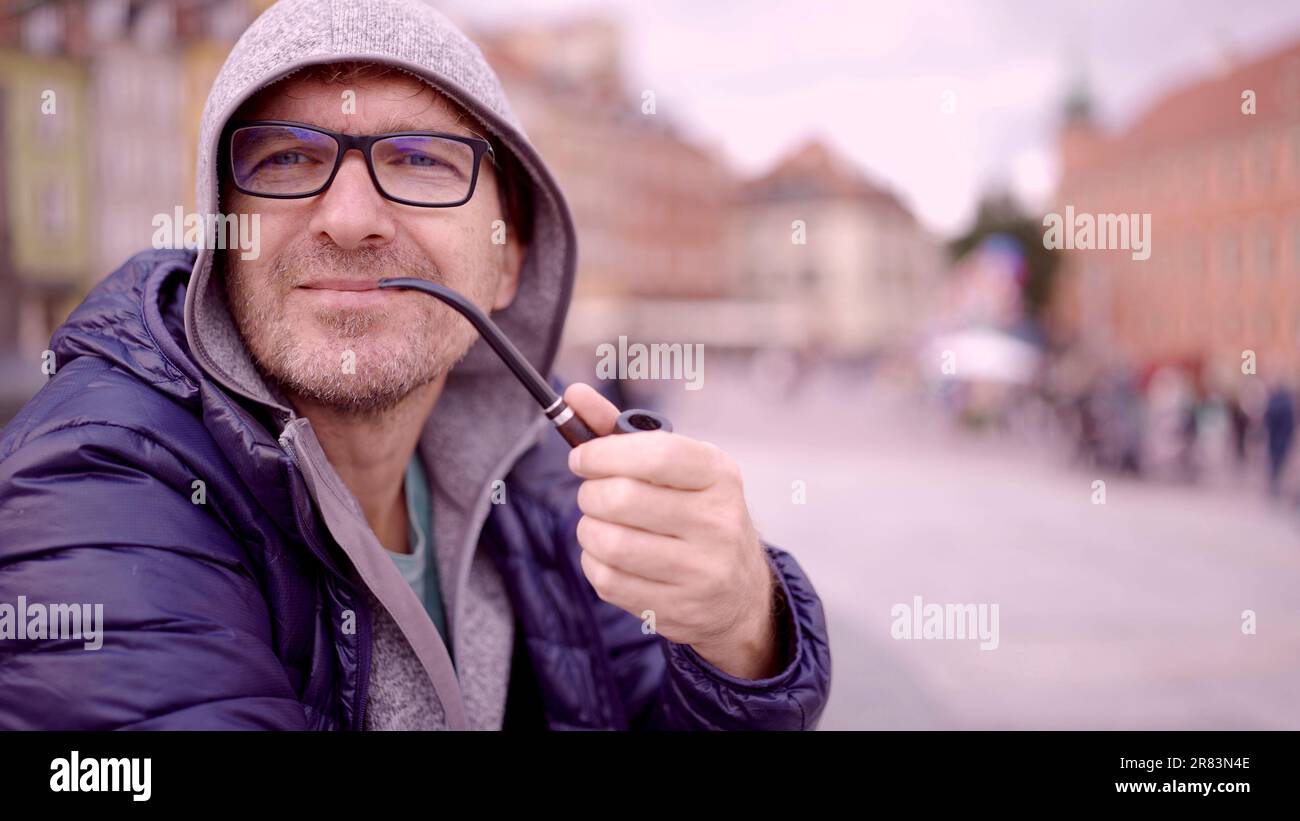 Porträt eines erwachsenen Mannes mit Brille, der in der Kapuze auf dem Platz sitzt und eine Tabakpfeife raucht, auf dem Palastplatz, Warschauer Altstadt Stockfoto