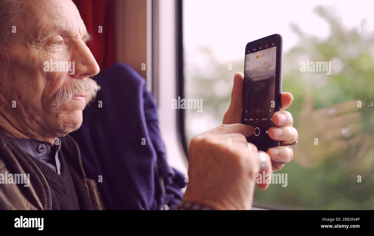 Älterer Mann, der mit dem Zug reist und mit einem Smartphone Bilder von der Landschaft durch das Fenster macht Stockfoto