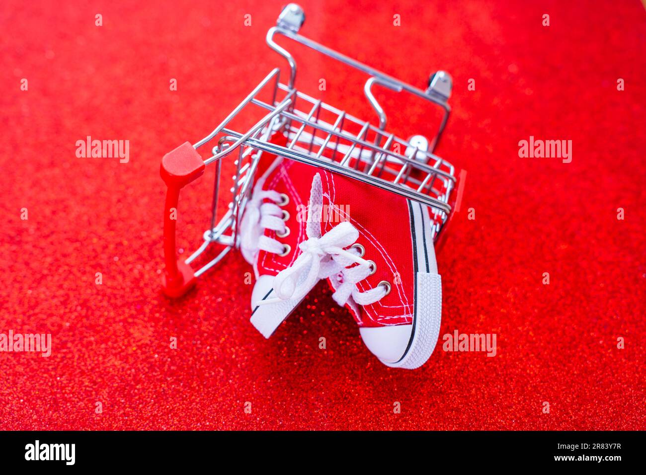 Ein Paar winzige rote Segelschuhe in einem umgedrehten Miniatur-Schubwagen vor einem glitzernden roten Hintergrund. Geschenke, Feiertage, E-Comm Stockfoto
