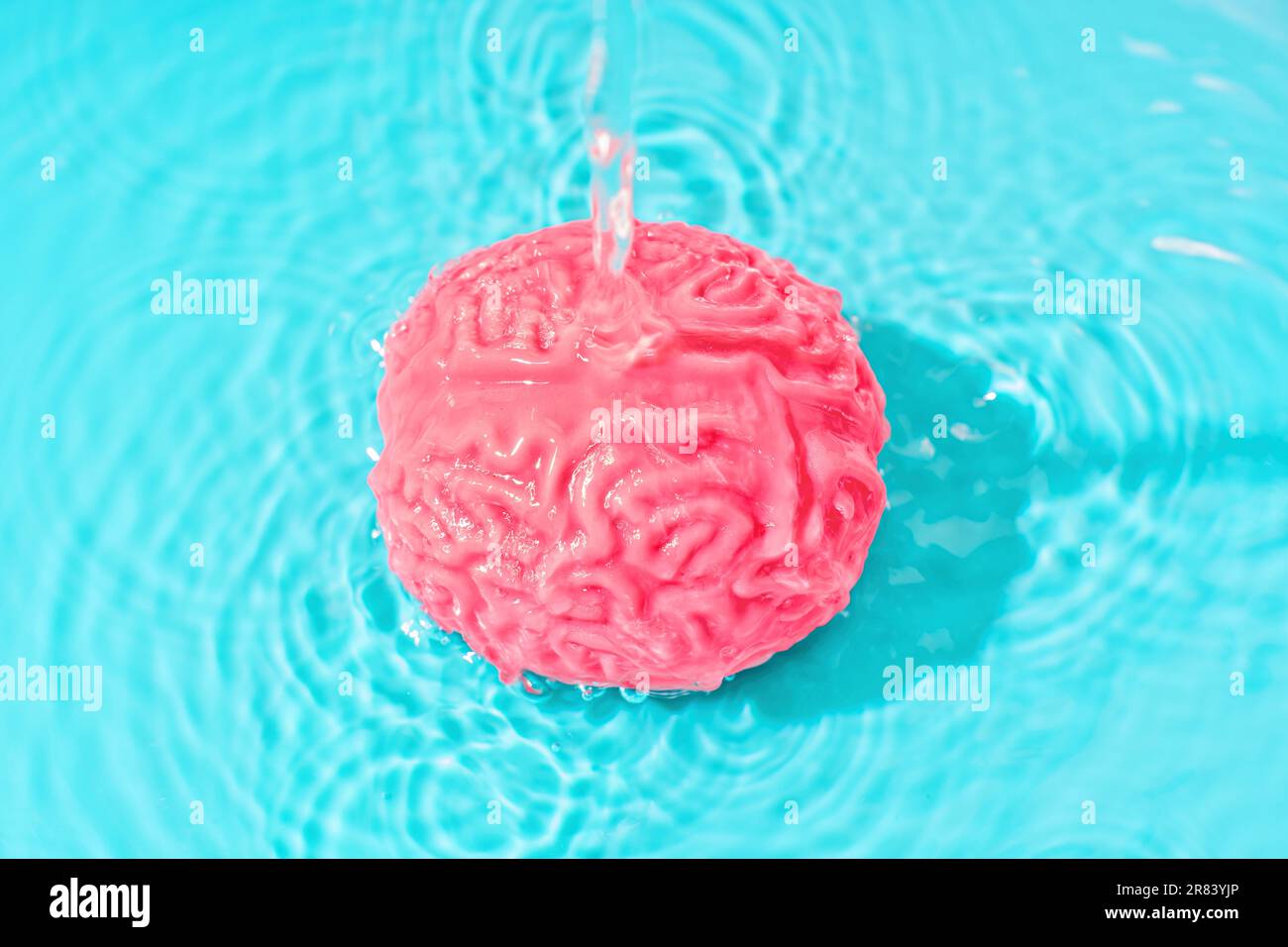 Ein stetiger Wasserstrom fließt auf ein gelartiges Modell des menschlichen Gehirns, das in Wasser vor einem ruhigen blauen Hintergrund eingetaucht ist. Manipulatio Stockfoto