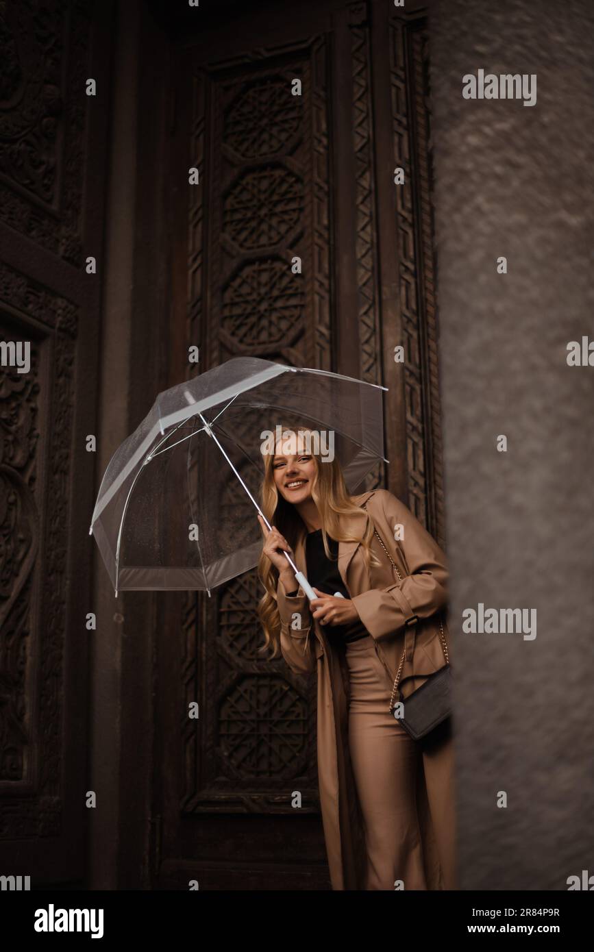 Glückliche, stilvolle Frau mit transparentem Regenschirm, die sich im alten Museumsgebäude der Stadt vor Regen versteckt Stockfoto