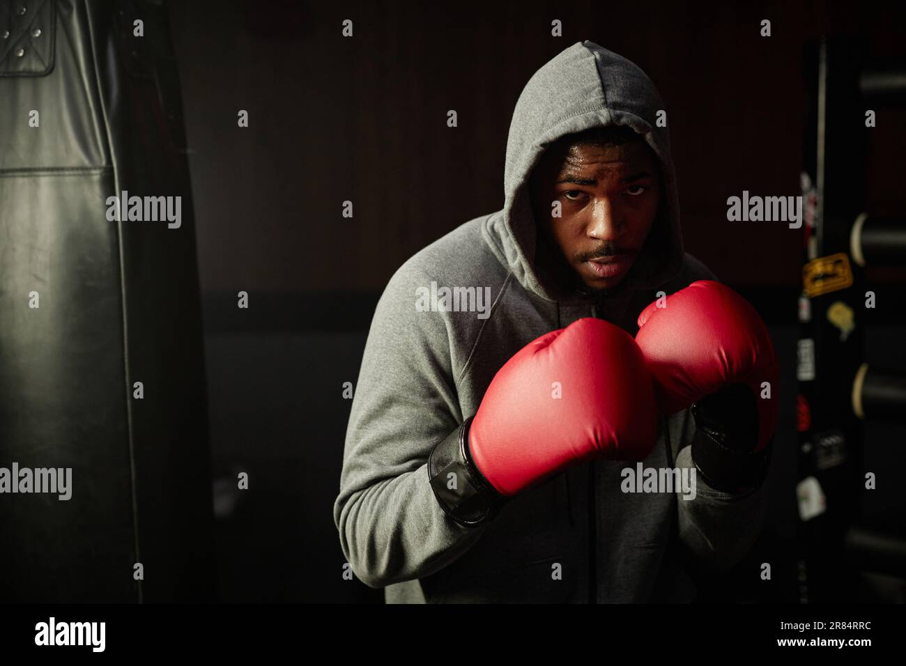 Junger, starker afroamerikanischer Sportler in roten Boxhandschuhen und grauem Hoodie, der auf die Kamera oder die Boxtasche schaut und sich auf den Kick vorbereitet Stockfoto