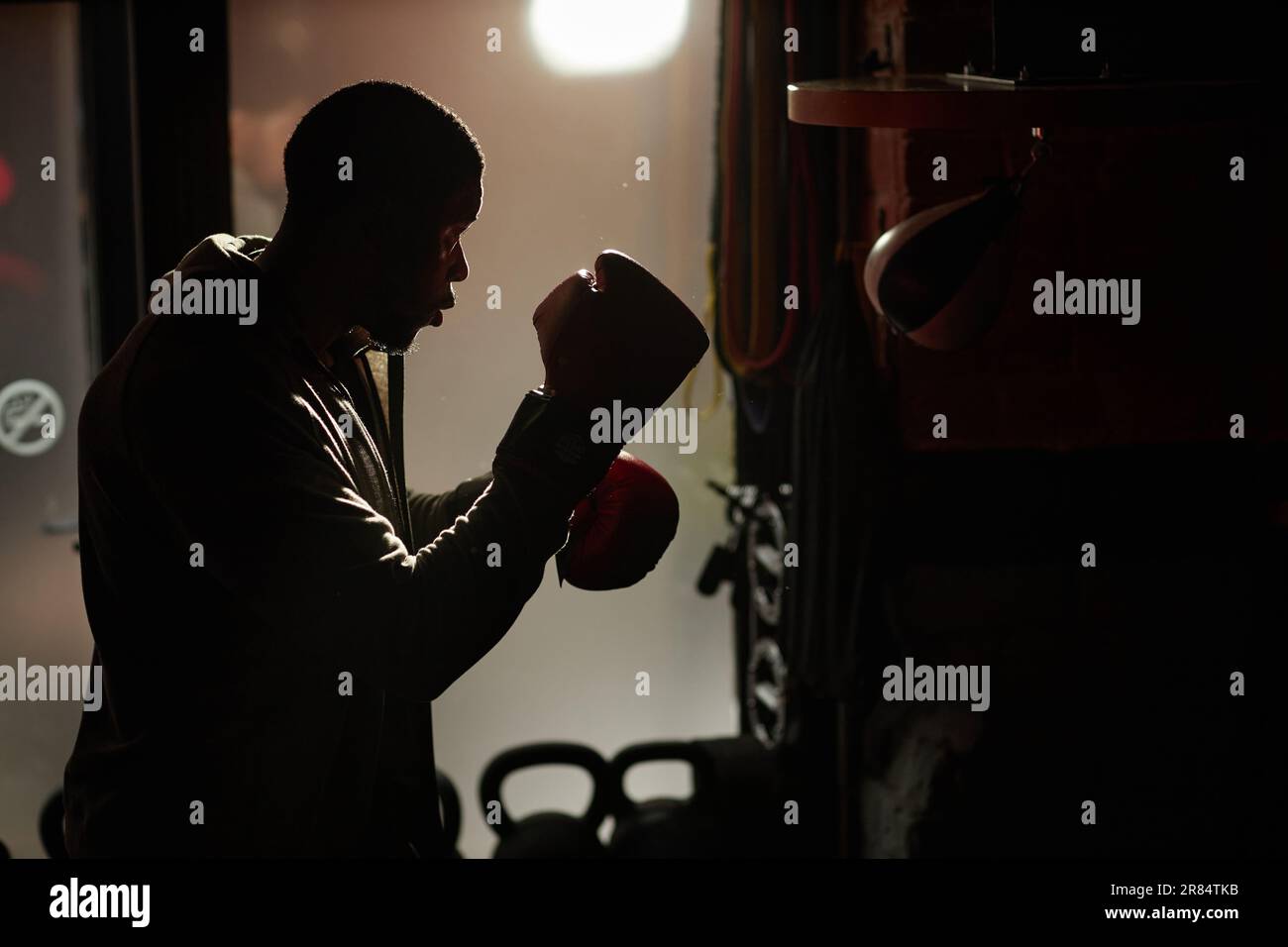 Entwurf eines jungen afroamerikanischen Boxers in Stanzhandschuhen, der im Fitnessstudio steht und beim Sporttraining auf einen Sandsack stößt Stockfoto