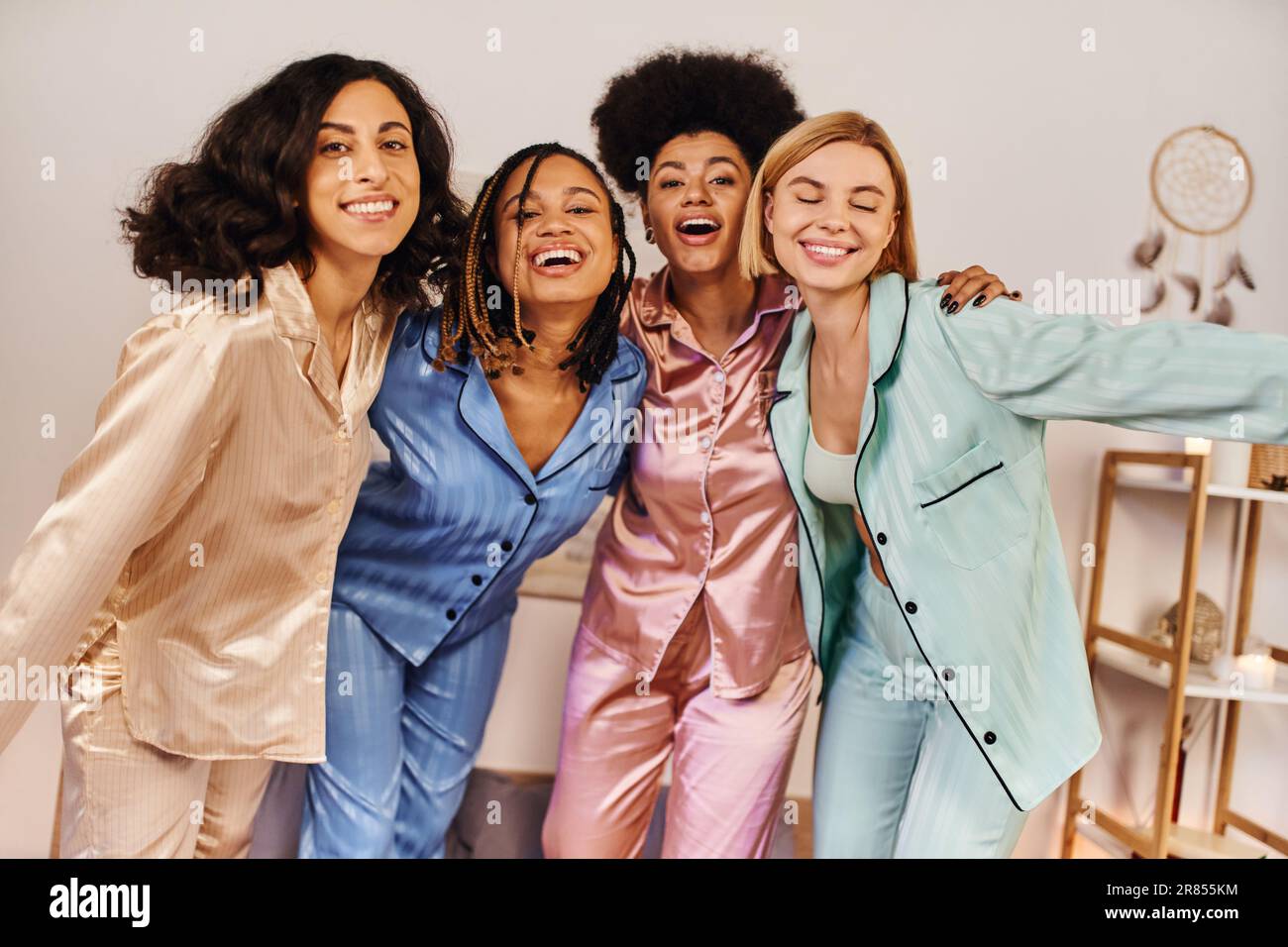 Lächelnde, multiethnische Freundinnen in einem bunten Pyjama, die in die Kamera schauen, sich umarmen und Spaß haben während der Pyjamaparty zu Hause, Zeit miteinander verbinden Stockfoto
