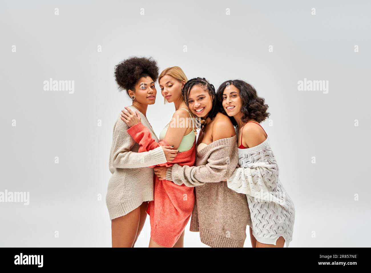 Fröhliche und stilvolle, multiethnische Frauen in Wollpullover, die sich umarmen und zusammenstehen, isoliert auf Grau, verschiedene Körpertypen und Selbstakzeptanz, mult Stockfoto