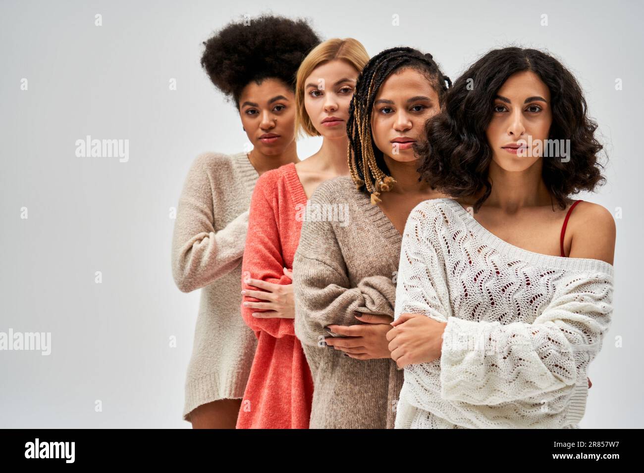Selbstbewusste und multiethnische Frauen in stilvollen Strickpullover, die die Arme kreuzen und nebeneinander stehen, isoliert auf grauen, verschiedenen Körpertypen und en Stockfoto