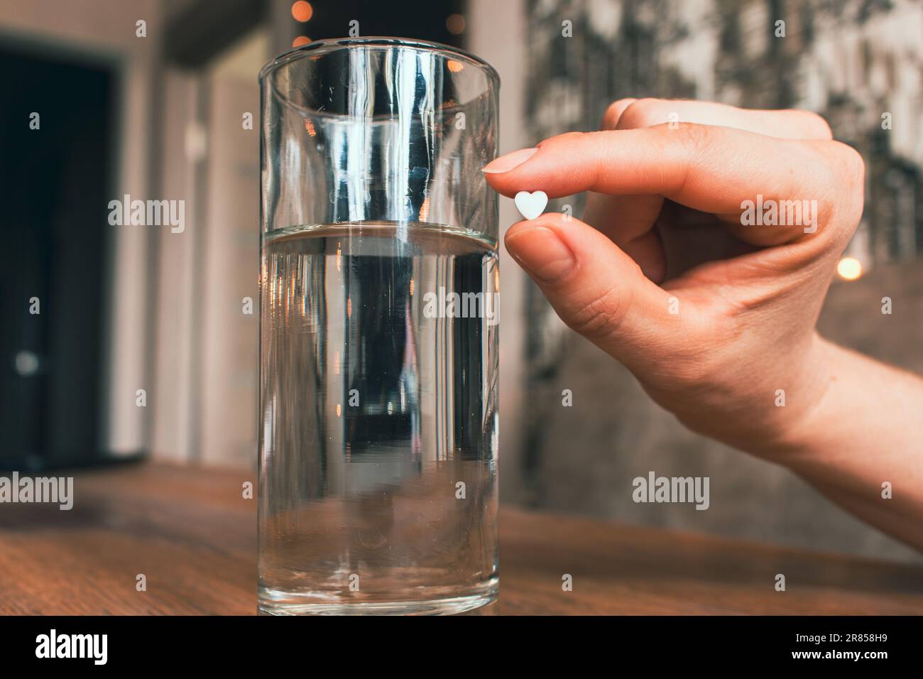 Weiße Pille in der Hand und ein Glas Wasser. Herzförmige Pille in der Hand. Medizinische Behandlung. Schmerzmittel aus unglücklicher Liebe. Therapierezeptur. Gesundheitswesen. Stockfoto