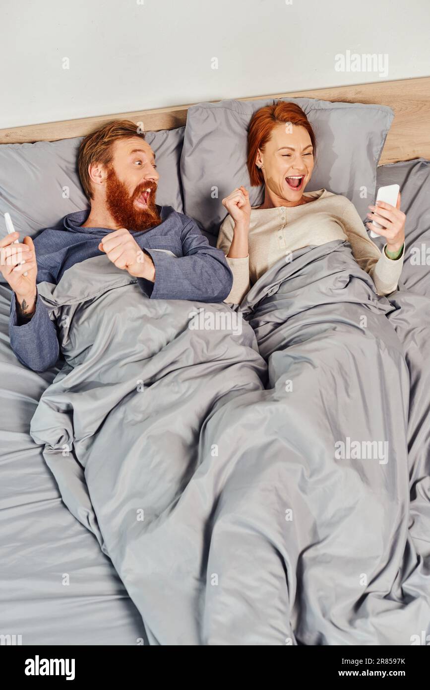 Tätowiertes Paar mit Smartphones, Networking, Entspannung am Wochenende ohne Kinder, aufgeregter Ehemann und Ehefrau, bärtiger Mann und rothaarige Frau mit Handy Stockfoto