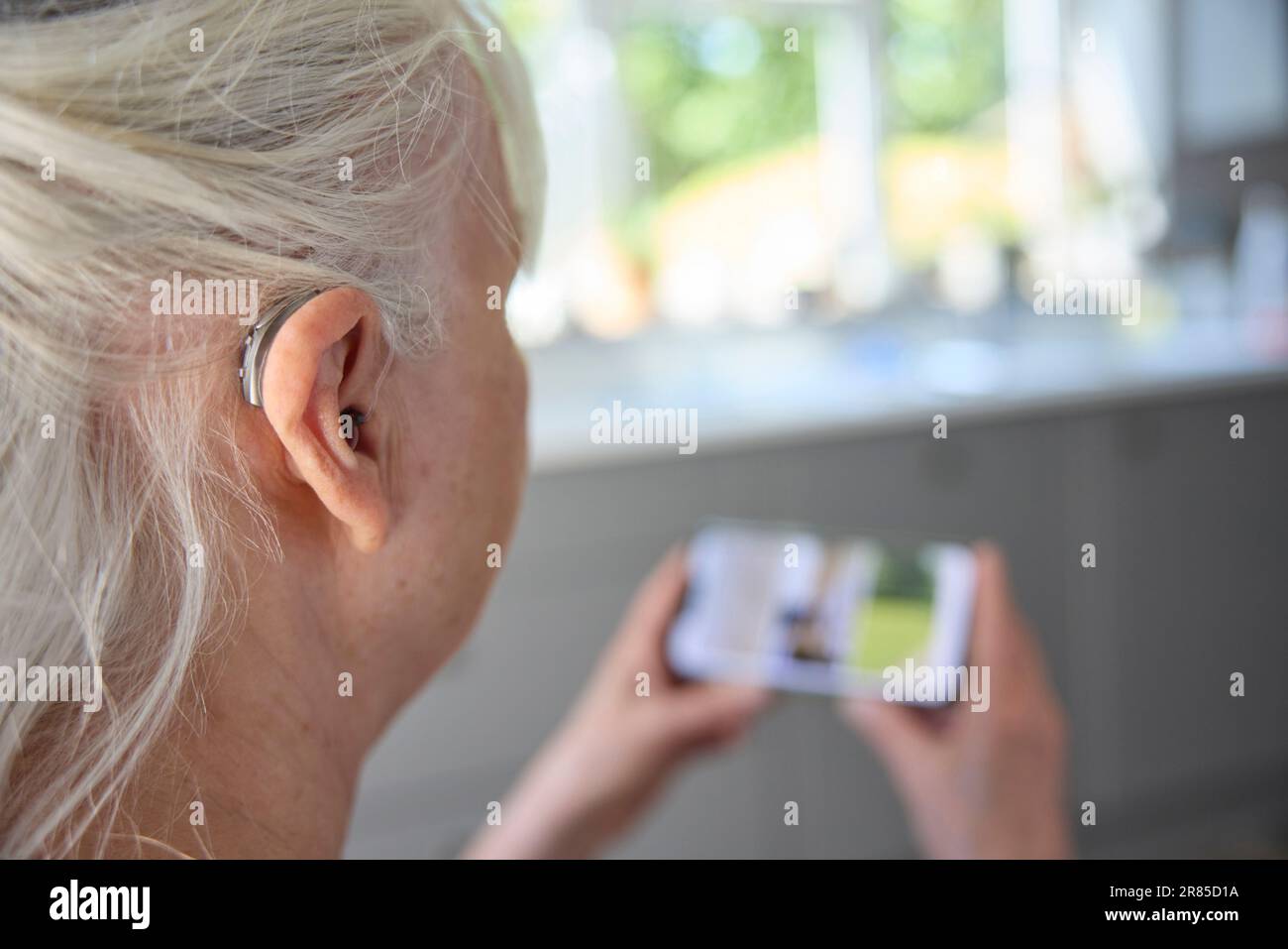 Nahaufnahme Einer Seniorin, Die Hinter Dem Ohr Ein Hörgerät Oder Ein Hörgerät Trägt, Das Zu Hause Über Ein Mobiltelefon Schleicht Stockfoto