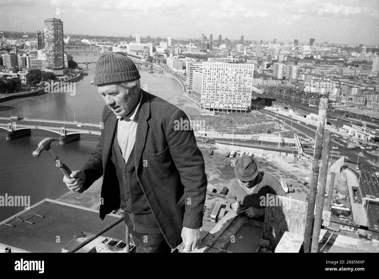 London Skyline 1970er. Irish Navvie Navvy ein irischer Arbeiter, ein Bauarbeiter, der einen Hammer an der Spitze hält und an einem Turmblock mit neun Elmen arbeitet. Blick auf die Londoner Stadtlandschaft, die Themse, Vauxhall Cross und Waterloo in der Ferne. London, England, ca. 1974. HOMER SYKES Stockfoto