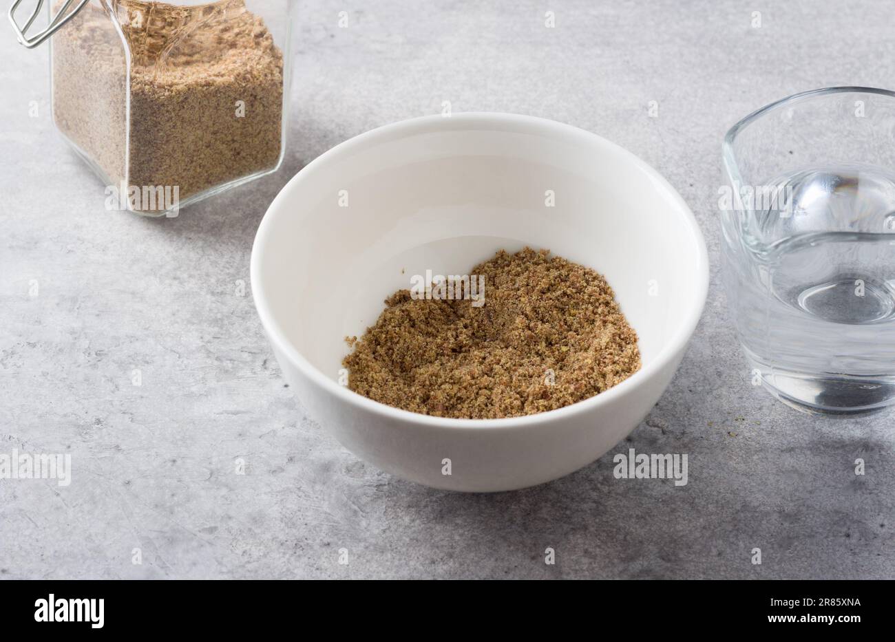 Eine Schüssel und ein Glas gemahlenen Flachs, eine Kanne Wasser. Veganes Flachsei auf grauem, strukturiertem Hintergrund kochen. Stockfoto