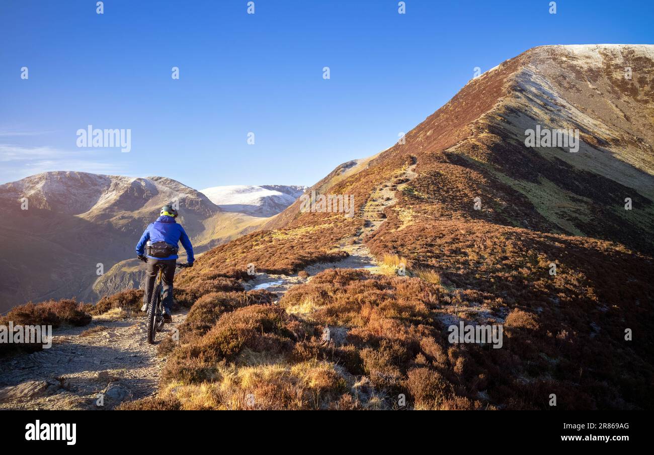 Ein Mountainbiker, der im Winter im englischen Lake District, Großbritannien, auf dem Weg zum Gipfel des Grisedale Pike mit Crag Hill in der Ferne reitet. Stockfoto