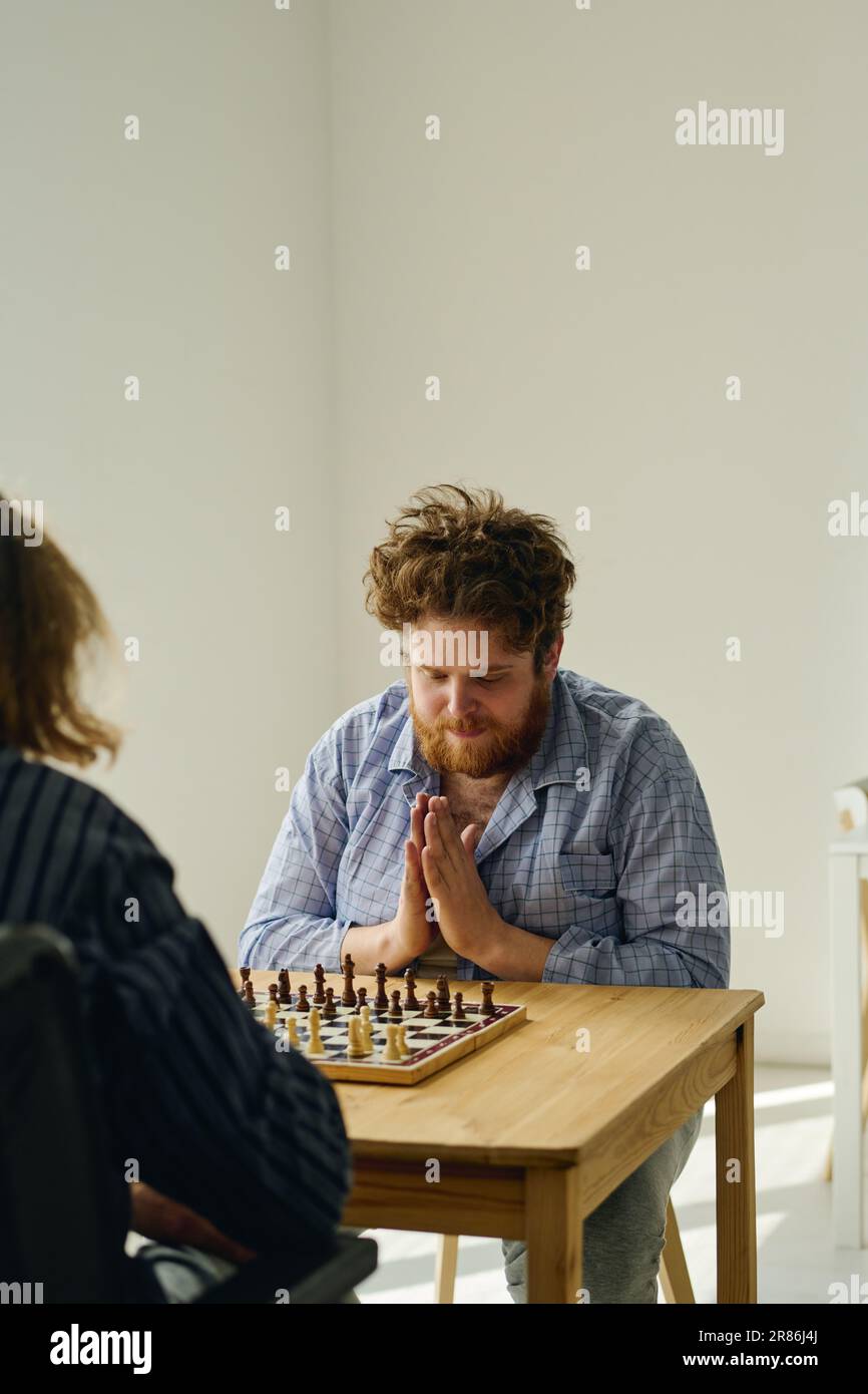 Junger bärtiger Mann, der die Hände an der Brust zusammenhält und auf das Schachbrett schaut, während er während des Spiels mit einem anderen Patienten an den nächsten Schritt denkt Stockfoto