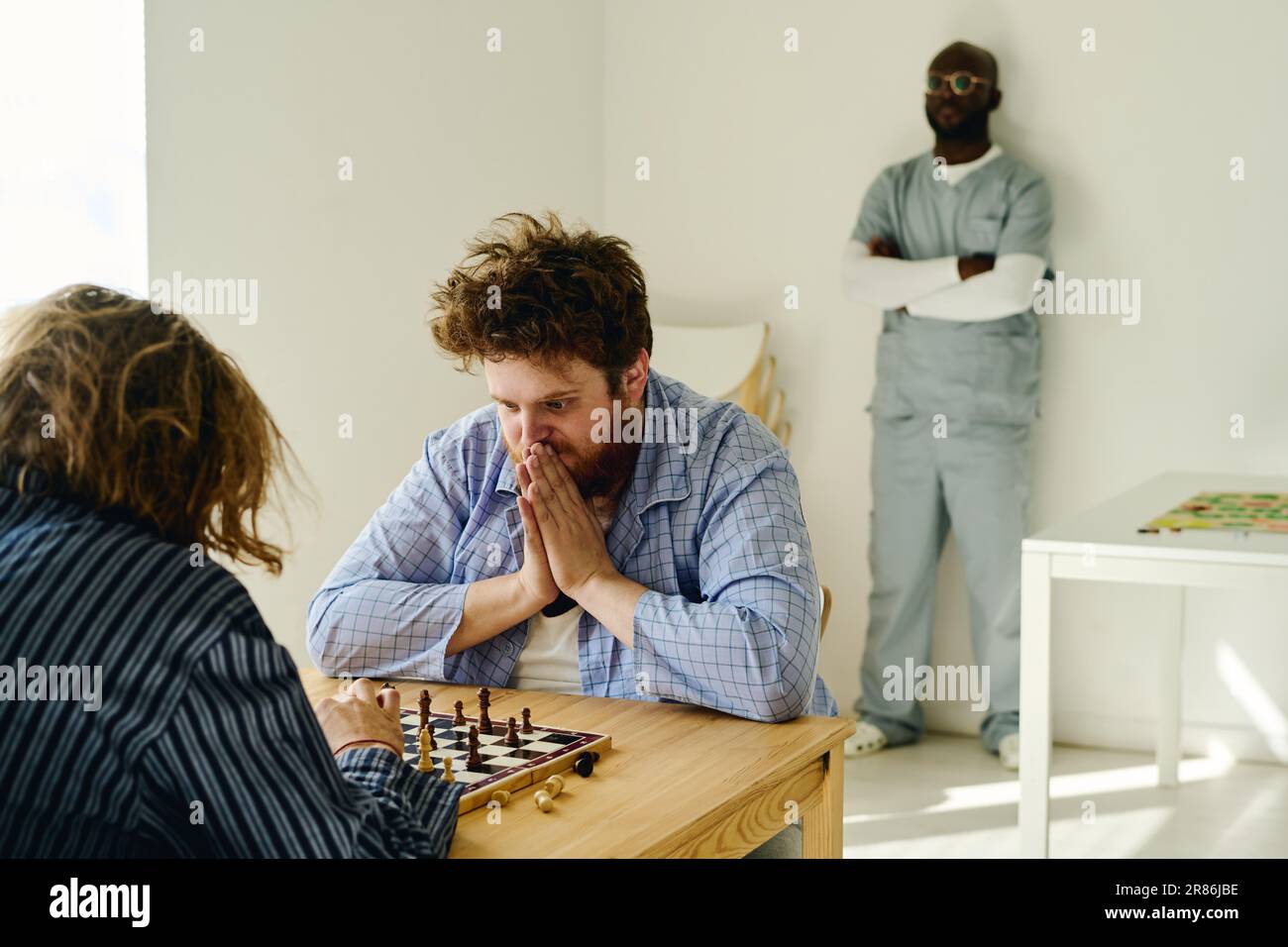 Junger nervöser Mann, der Handflächen von Gesicht zu Gesicht zusammenhält und Schachbrett betrachtet, während er mit einem anderen männlichen Patienten der Nervenklinik spielt Stockfoto