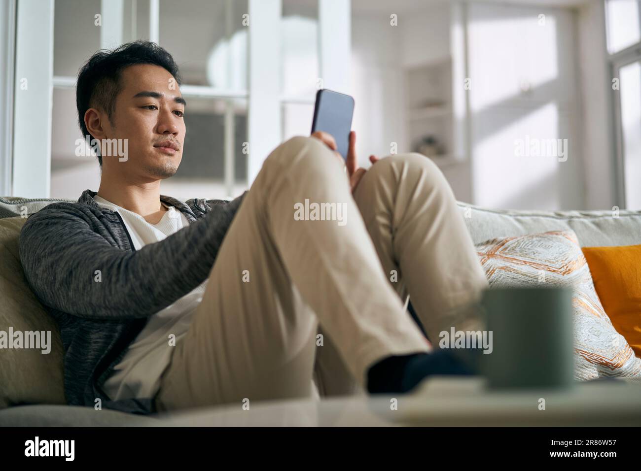 Junger asiatischer Erwachsener, der auf dem Familientisch im Wohnzimmer zu Hause sitzt und auf das Handy schaut Stockfoto