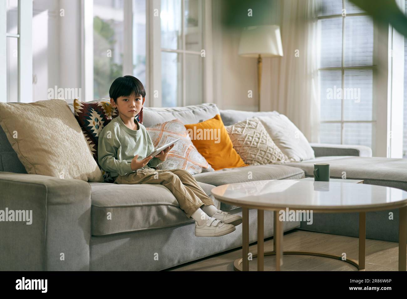 Kleiner asiatischer Junge, der auf der Familientischcouch sitzt, im Wohnzimmer und in die Kamera schaut Stockfoto