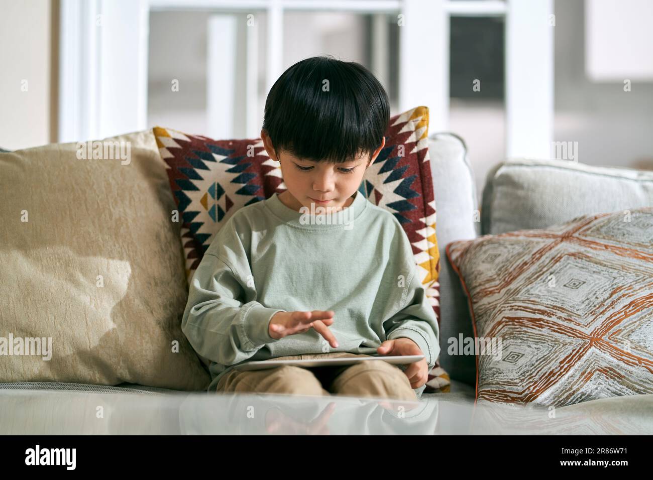 Fünf Jahre alter kleiner asiatischer Junge, der zu Hause auf dem Familientisch sitzt und einen digitalen Tablet-Computer benutzt Stockfoto
