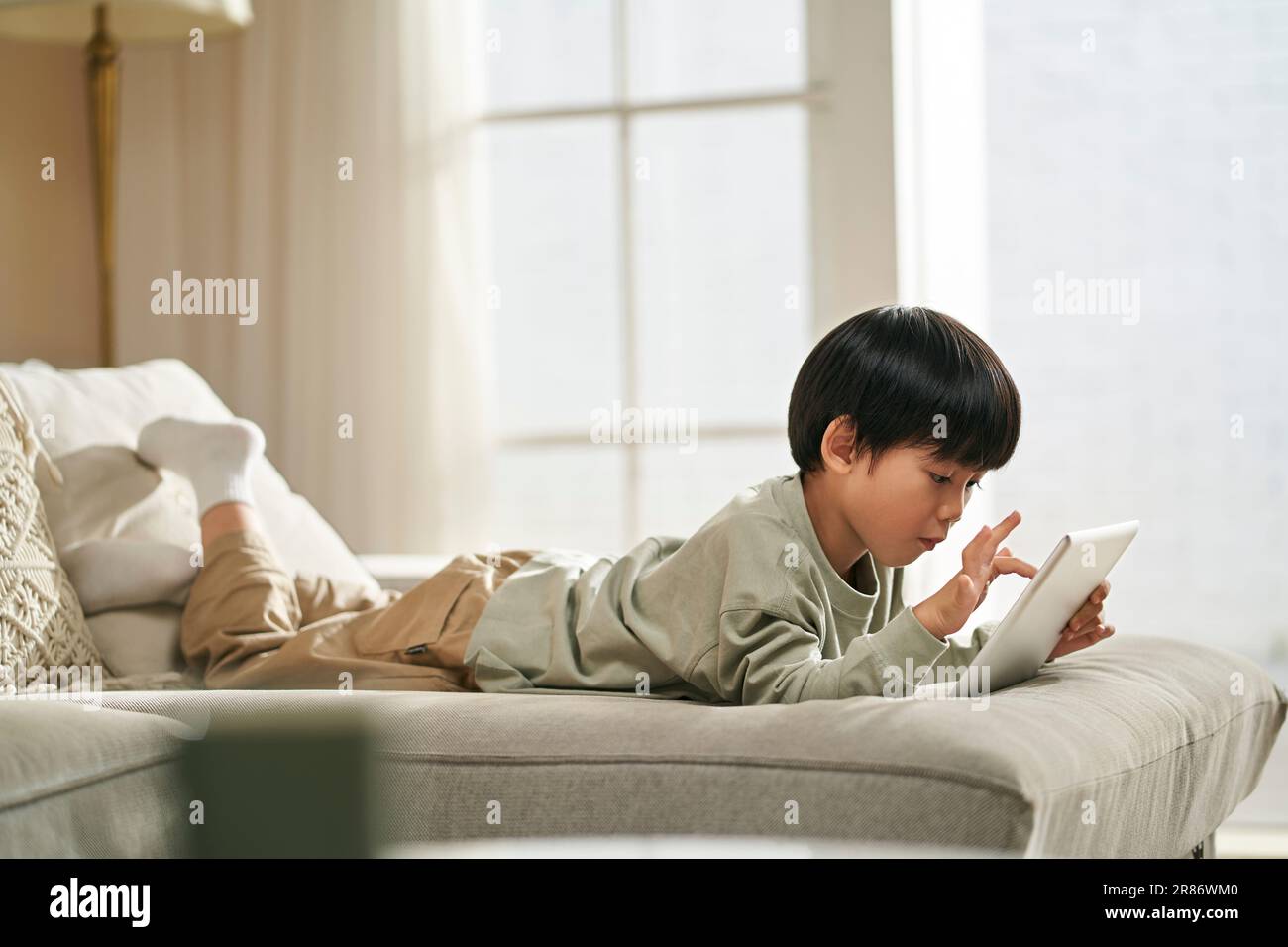 Fünf Jahre alter kleiner asiatischer Junge, der vorne auf dem Familientisch liegt und Computerspiel mit einem digitalen Tablet spielt Stockfoto
