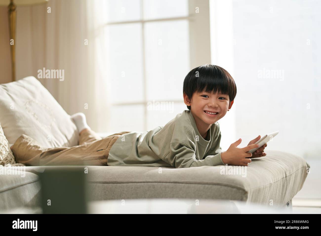Süßer kleiner asiatischer Junge, der zu Hause vorne auf dem Familientisch liegt und ein digitales Tablet hält Stockfoto