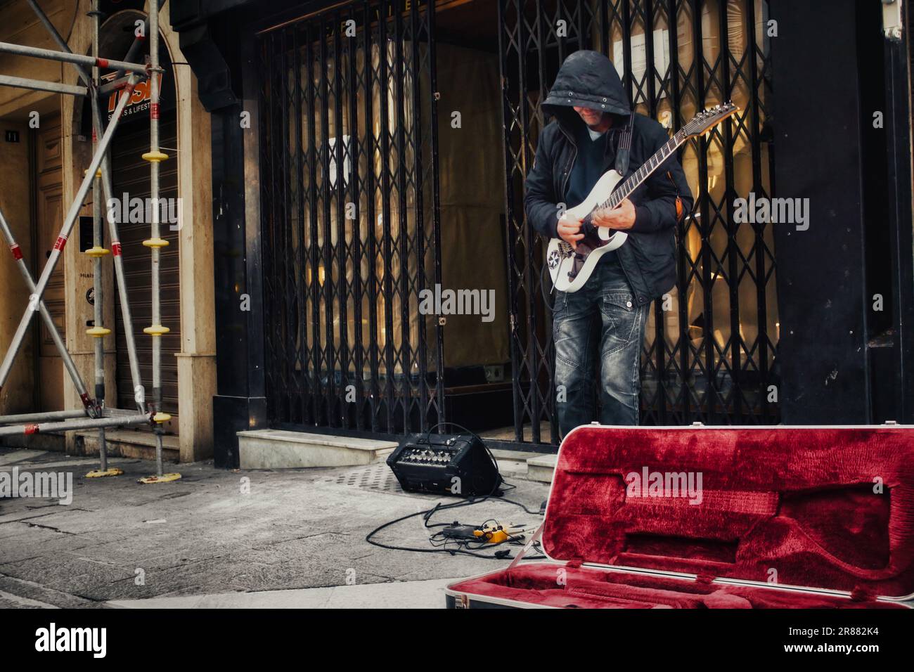 Ein unbekannter, mit Kapuze besetzter Straßenmusiker in einer Fußgängerzone im Stadtzentrum, der eine elektrische Blues-Gitarre mit Verstärker und Pedalen spielt Stockfoto