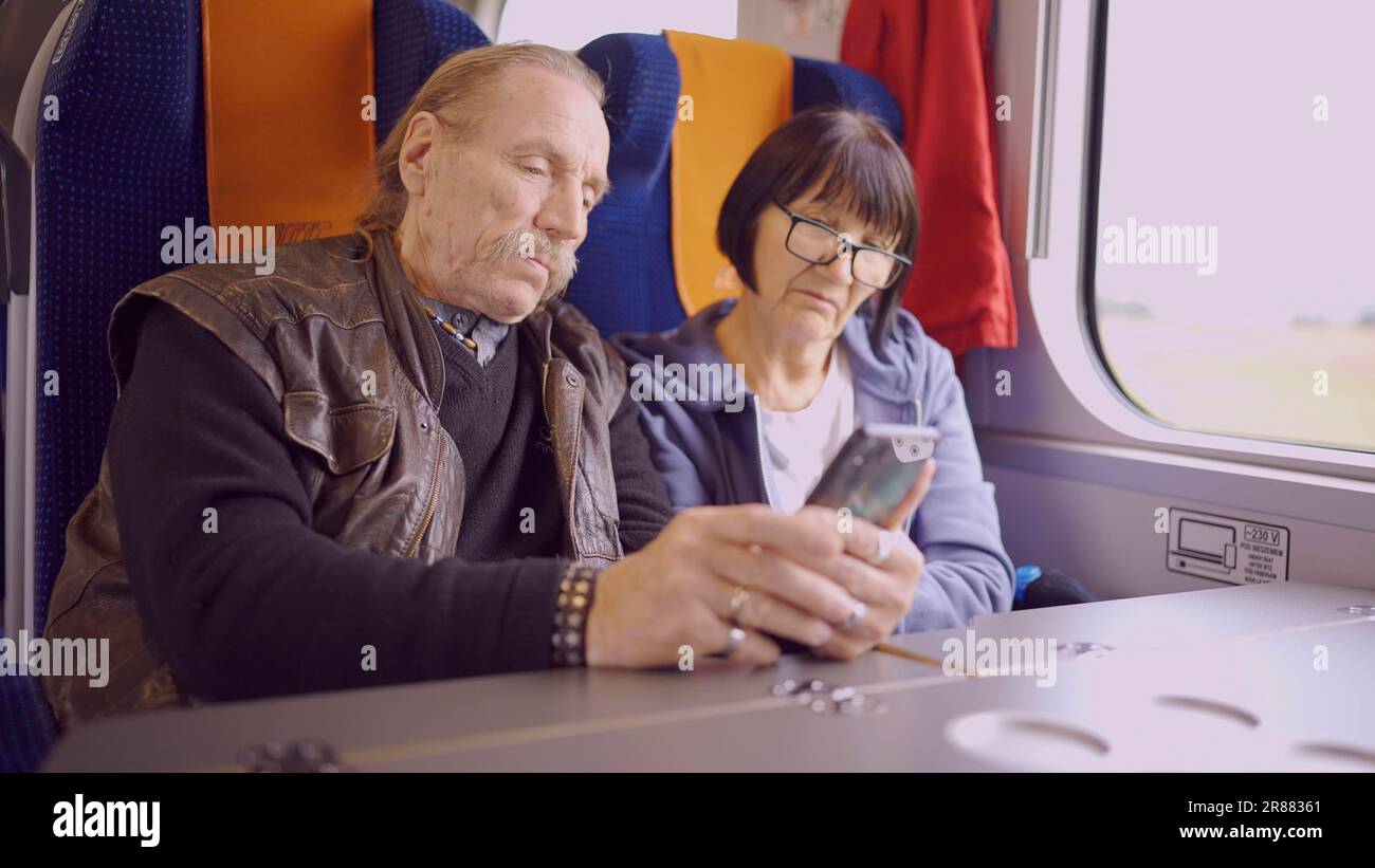 Ältere Paare reisen mit dem Zug, die Seniorin hält ein Handy in der Hand, beide schauen auf das Smartphone und sprechen miteinander Stockfoto