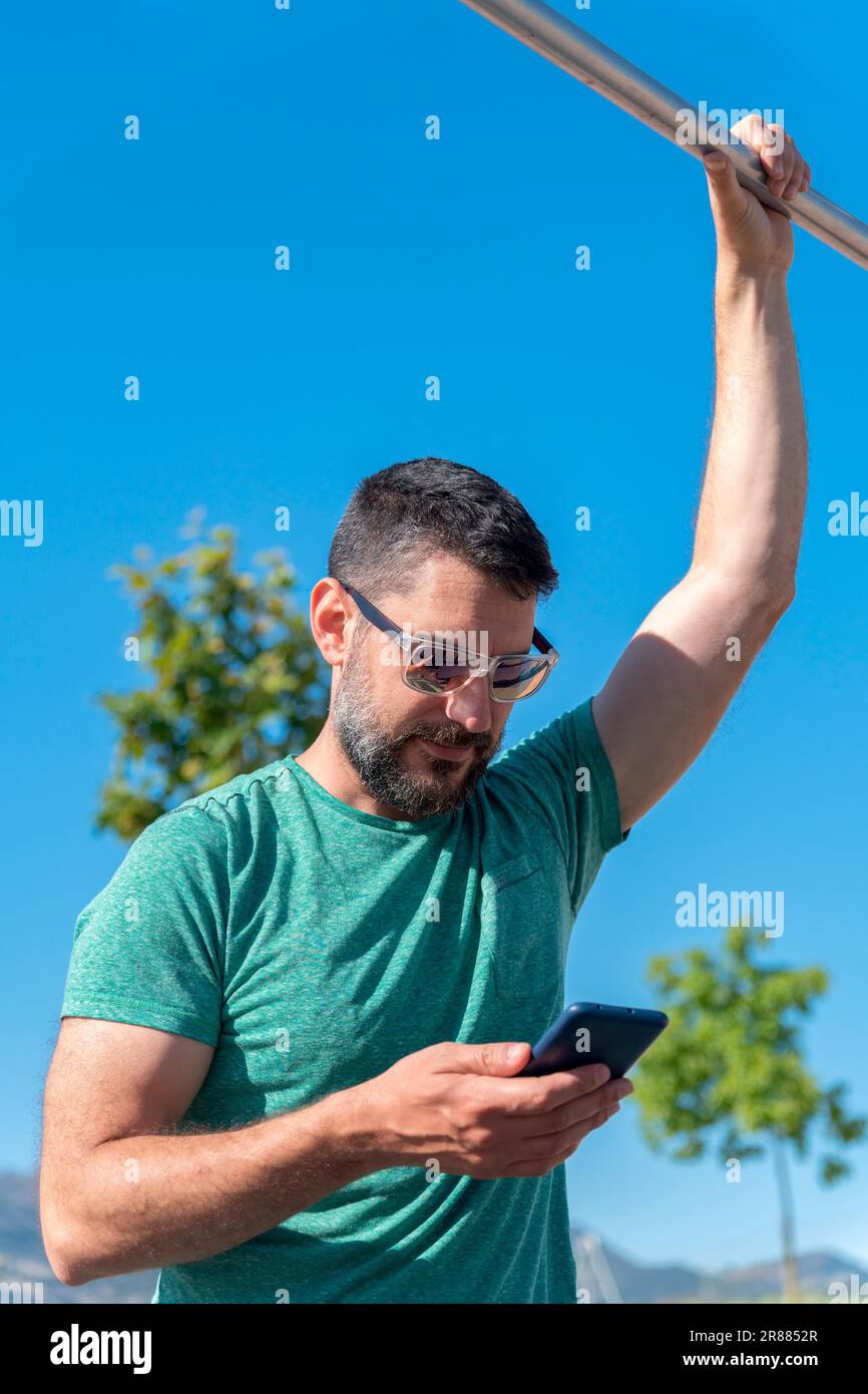 Ein junger Mann mit Bart und Sonnenbrille konsultiert eine Trainingsanwendung auf seinem Mobiltelefon, während er in einem Fitnessstudio im Park trainiert Stockfoto