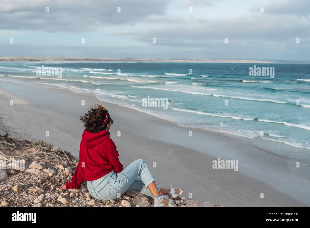Eine Frau mit kurzen Haaren und rotem Kapuzenpulli, die an einem felsigen Strand mit Blick auf das Meer sitzt Stockfoto