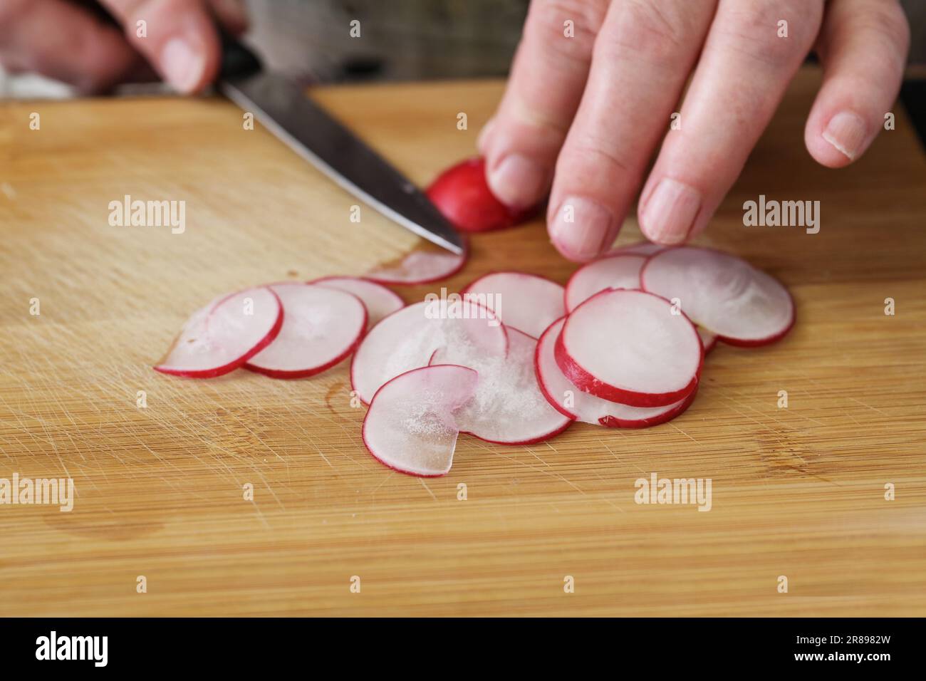 Die Hände schneiden Radieschen mit einem Küchenmesser auf einem Holzbrett in dünne Scheiben für einen frischen Salat, Kopierraum, ausgewählten Fokus, geringe Falltiefe Stockfoto